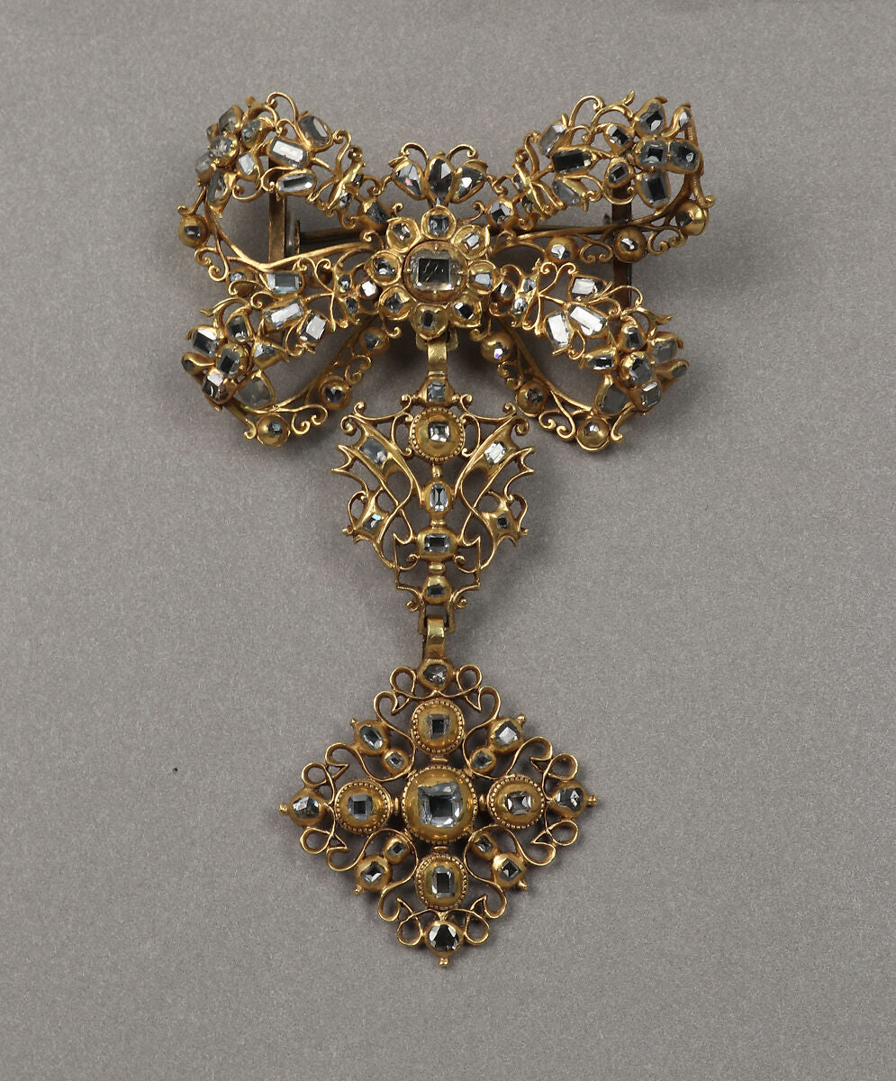An ornate openwork bow with a central floral setting and an articulated decorative drop. Fine gold scrolls surround the dark reflective diamond settings.
